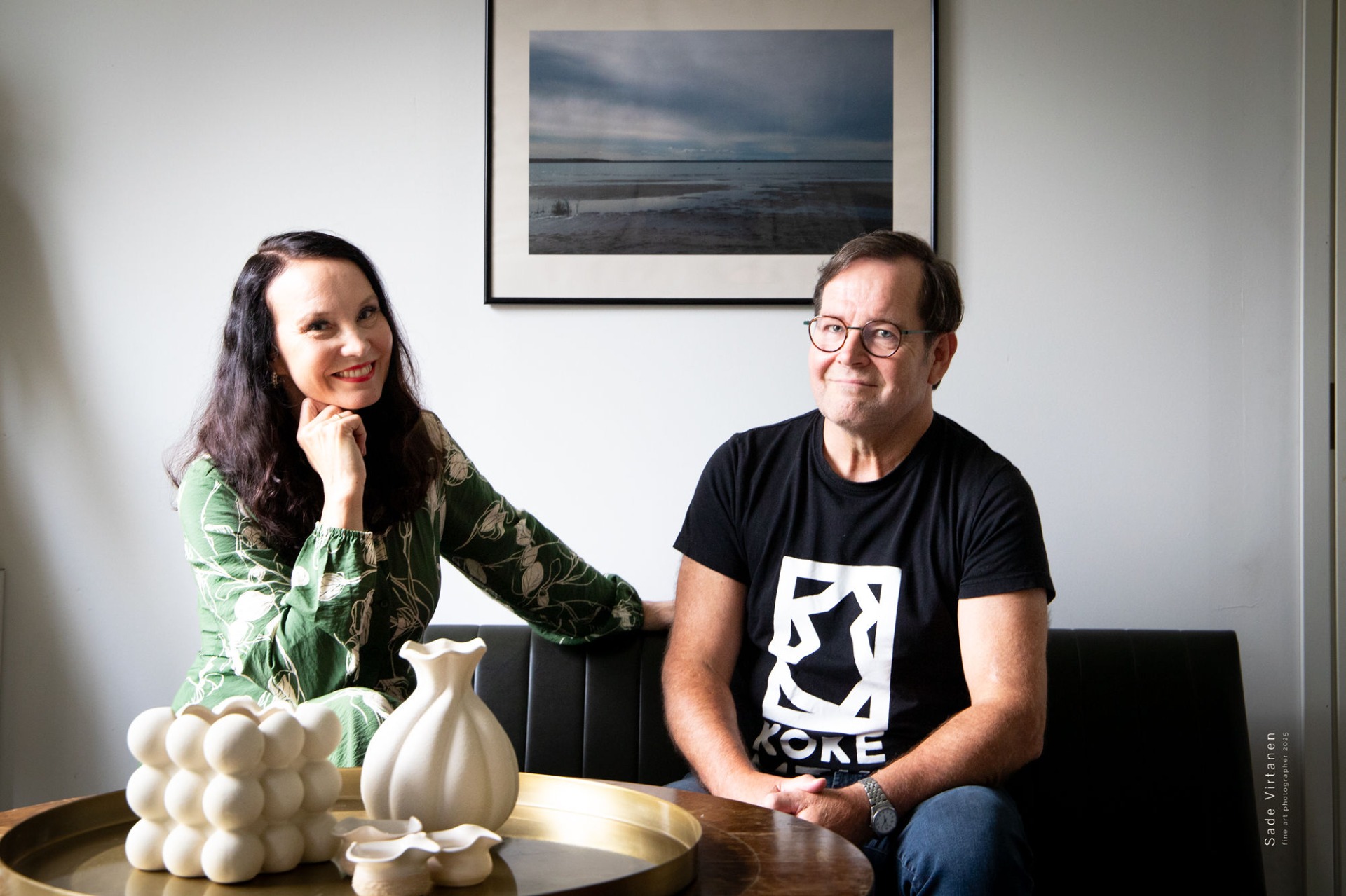 Johanna Rantalainen ja Juhani Seppälä istuvat sohvalla valokuvaamon galleriassa, taustalla maisemataulu.