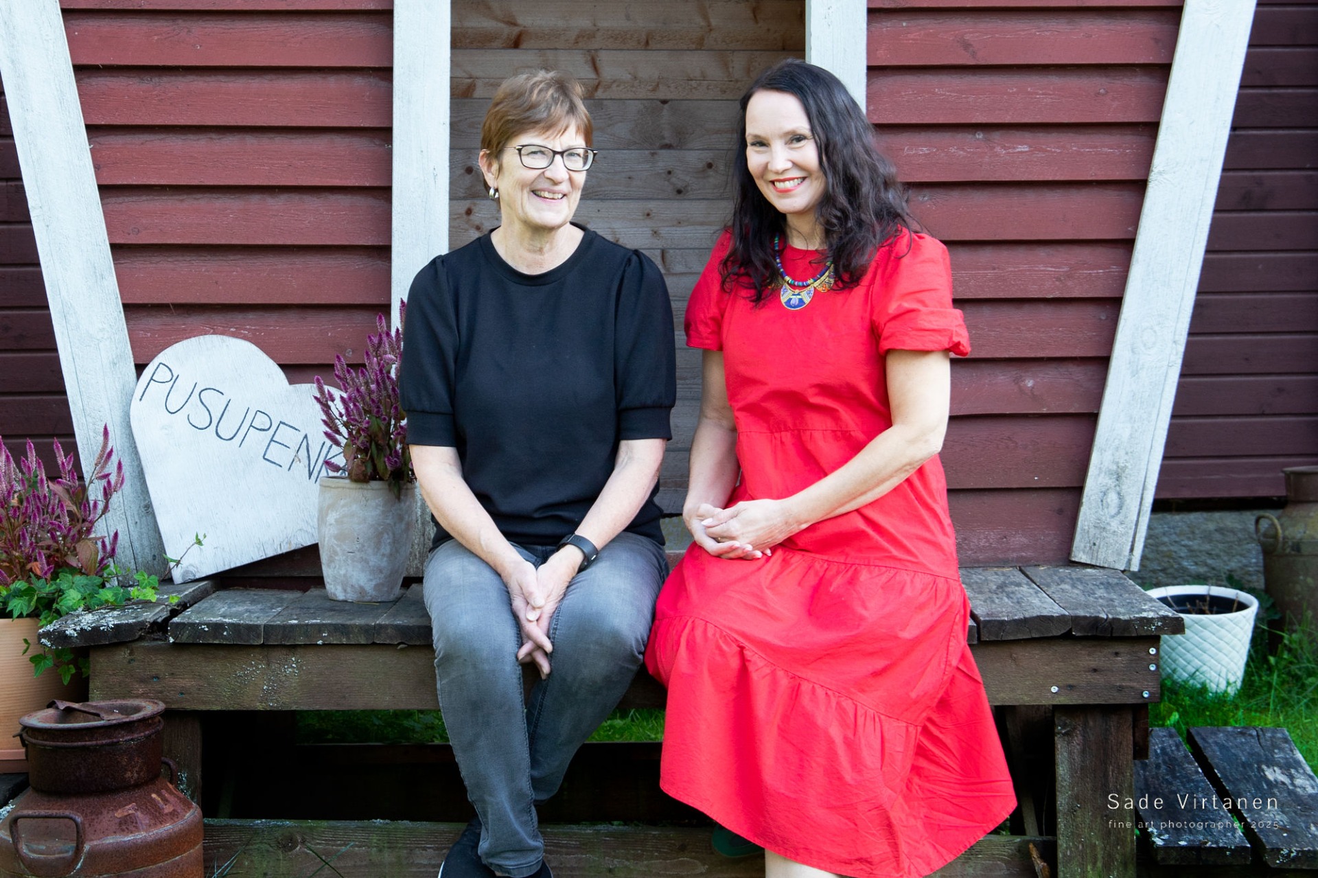 Yrittäjä Sirpa Ala-Ketola ja Johanna Rantalainen istuvat  punaisella maitolaiturilla. Sirpalla on päällään musta T-paita ja farkut. Johannalla on punainen mekko.